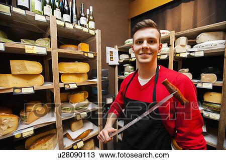 Stock Image - Portrait of confident male salesperson holding knife in cheese store. Fotosearch