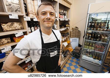 Stock Photograph - Portrait of confident mid adult salesman standing hands on hips in grocery store. Fotosearch