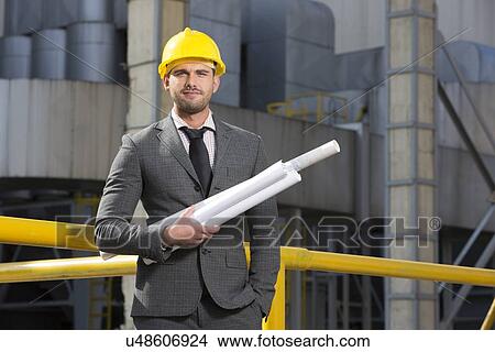 Portrait of confident young architect holding blueprints outside industry View Large Photo Image Picture - Portrait of confident young architect holding blueprints outside industry. Fotosearch