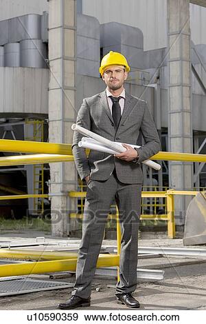 Stock Photo - Portrait of confident young businessman holding rolled up blueprints outside industry. Fotosearch