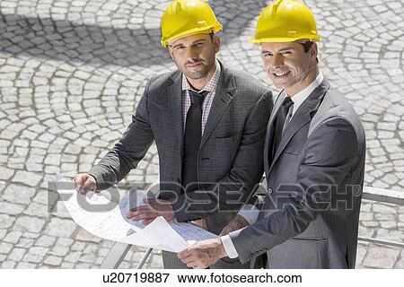 Stock Photo - Portrait of confident young businessmen in hard hats examining blueprint outdoors. Fotosearch