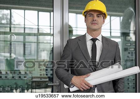 Portrait of confident young male architect holding rolled up blueprints in industry View Large Photo Image Stock Photo - Portrait of confident young male architect holding rolled up blueprints in industry. Fotosearch