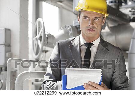 Stock Photo - Portrait of confident young male supervisor writing on clipboard in industry. Fotosearch