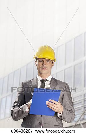 Stock Photo - Portrait of confident young male architect writing on clipboard outside building. Fotosearch