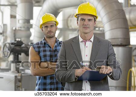 Picture - Portrait of confident young manager writing on clipboard with manual worker in background at industry. Fotosearch