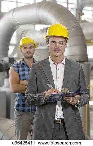 Stock Image - Portrait of confident young manager writing on clipboard with manual worker in background at industry. Fotosearch