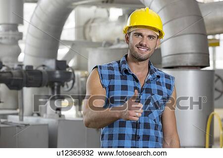 Stock Image - Portrait of confident young worker gesturing thumbs up in industry. Fotosearch