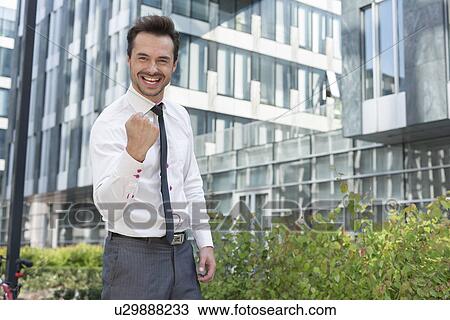 Stock Image - Portrait of happy businessman with clenched fist standing outside office building. Fotosearch