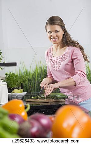 Stock Image - Portrait of happy woman cutting cucumber at kitchen counter. Fotosearch