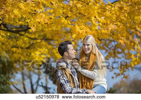 Stock Photo - Portrait of happy woman sitting on man's lap in park during autumn. Fotosearch