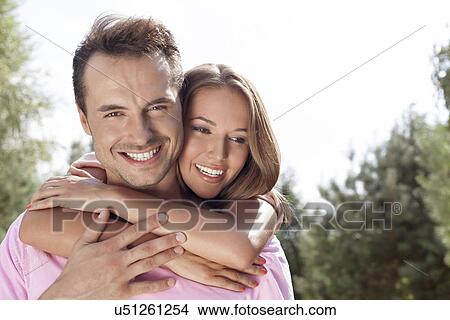 Picture - Portrait of happy young man being embrace by woman from behind in park. Fotosearch