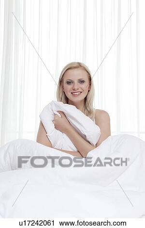 Stock Image - Portrait of happy young woman holding bedsheet in bed. Fotosearch