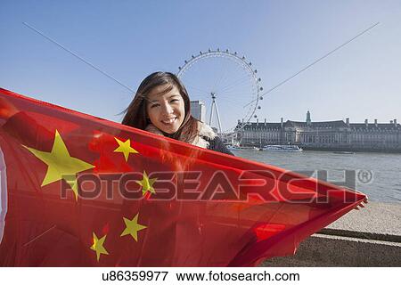 Stock Photo - Portrait of happy young woman holding Chinese flag against London Eye at London, England, UK. Fotosearch
