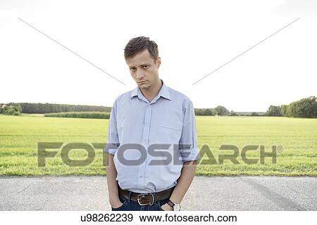 Stock Photo - Portrait of sad young man with hands in pockets standing on rural road. Fotosearch