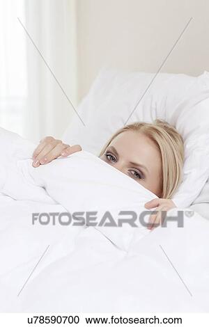 Stock Image - Portrait of shy young woman hiding under bedsheet. Fotosearch