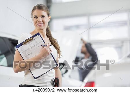 Stock Image - Portrait of smiling female repair worker with clipboard in car workshop. Fotosearch