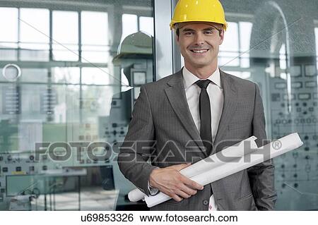 Portrait of smiling young architect holding rolled up blueprints in industry View Large Photo Image Stock Photograph - Portrait of smiling young architect holding rolled up blueprints in industry. Fotosearch