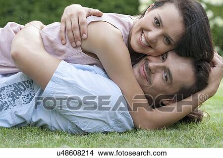 Picture - Portrait of smiling young couple embracing while lying in park. Fotosearch