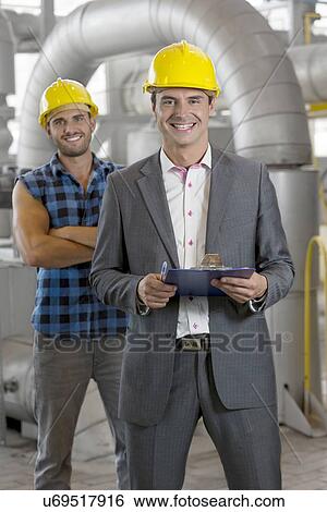 Stock Photograph - Portrait of smiling young male manager holding clipboard with worker in background at industry. Fotosearch