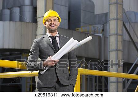 Stock Image - Portrait of smiling young male architect holding blueprints outside building. Fotosearch