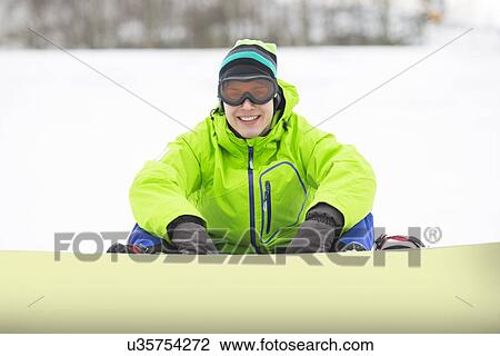 Stock Image - Portrait of smiling young man with snowboard sitting on snowy land. Fotosearch