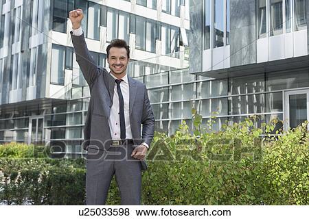 Stock Photo - Portrait of successful businessman with arm raised standing outside office building. Fotosearch