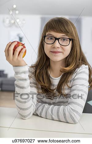 Stock Photo - Portrait of teenage girl holding apple while sitting at table in house. Fotosearch