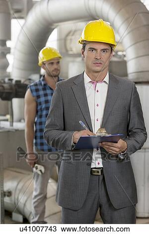 Portrait of young male manager writing on clipboard with manual worker in background at industry View Large Photo Image Stock Image - Portrait of young male manager writing on clipboard with manual worker in background at industry. Fotosearch