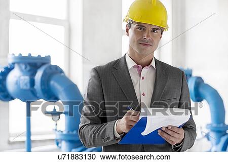 Stock Image - Portrait of young male manager writing on clipboard in industry. Fotosearch
