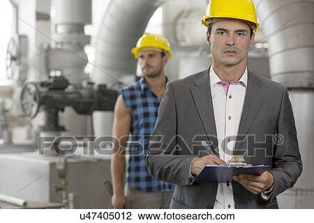 Portrait of young male supervisor writing on clipboard with manual worker in background at industry View Large Photo Image Stock Image - Portrait of young male supervisor writing on clipboard with manual worker in background at industry. Fotosearch