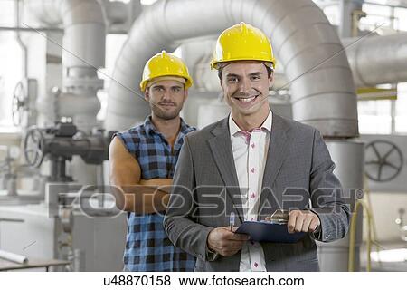 Portrait of young male supervisor holding clipboard with manual worker in background at industry View Large Photo Image Stock Photo - Portrait of young male supervisor holding clipboard with manual worker in background at industry. Fotosearch