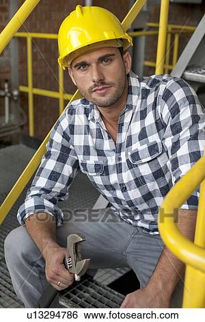 Stock Photograph - Portrait of young male worker with wrench sitting on steps at industry. Fotosearch