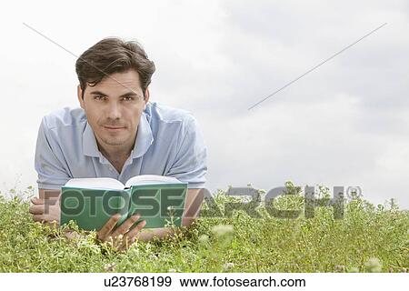 Stock Photo - Portrait of young man holding book while lying on grass against sky. Fotosearch
