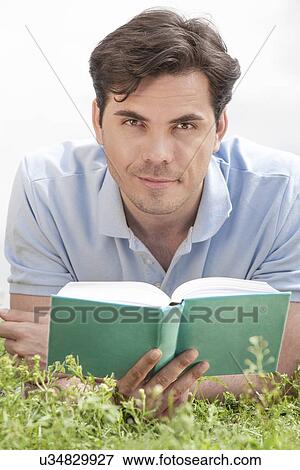 Portrait of young man holding book while lying on grass View Large Photo Image Stock Photo - Portrait of young man holding book while lying on grass. Fotosearch