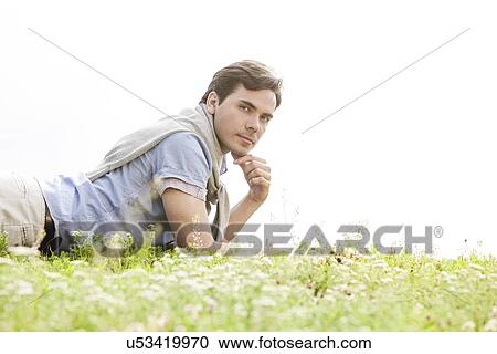 Stock Image - Portrait of young man lying on grass against clear sky. Fotosearch