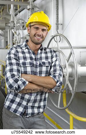 Stock Image - Portrait of young worker with arms crossed leaning on large valve in industry. Fotosearch
