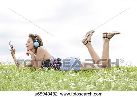 Stock Image - Profile shot of woman listening to music through cell phone using headphones while lying on grass against sky. Fotosearch