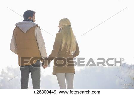 Stock Photo - Rear view of couple holding hands while looking at each other in park. Fotosearch