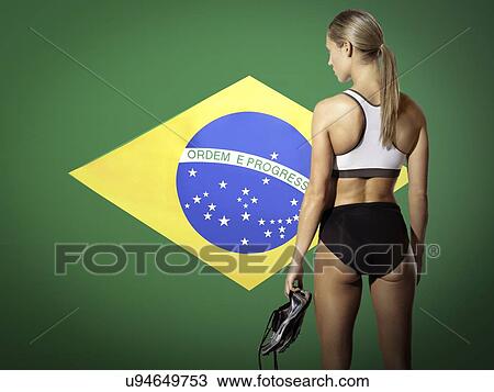 Stock Image - Rear view of female runner holding shoes while standing against Brazilian flag. Fotosearch