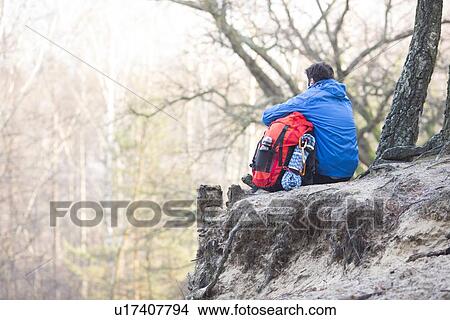 Rear view of hiker with backpack sitting on edge of cliff in forest View Large Photo Image Picture - Rear view of hiker with backpack sitting on edge of cliff in forest. Fotosearch