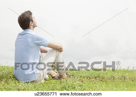 Rear view of relaxed young man sitting on grass against sky View Large Photo Image Stock Image - Rear view of relaxed young man sitting on grass against sky. Fotosearch