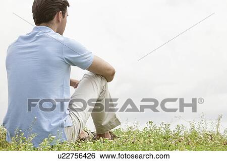 Stock Photograph - Rear view of young man sitting on grass against sky. Fotosearch