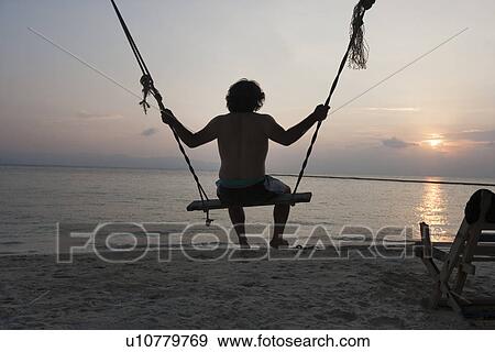 Rear view of young man swinging on beach at sunset View Large Photo Image Stock Photo - Rear view of young man swinging on beach at sunset. Fotosearch