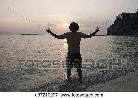 Rear view of young man with arms outstretched on beach at sunset View Large Photo Image Stock Photo - Rear view of young man with arms outstretched on beach at sunset. Fotosearch