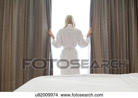 Picture - Rear view of young woman in bathrobe opening window curtains at hotel room. Fotosearch