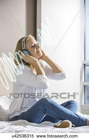 Relaxed mid adult woman listening music through headphones on bed View Large Photo Image Stock Photography - Relaxed mid adult woman listening music through headphones on bed. Fotosearch