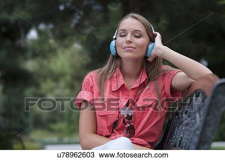 Stock Image - Relaxed young woman listening to headphones on park bench. Fotosearch