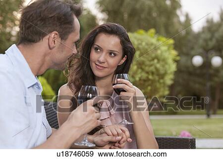 Stock Photo - Romantic young couple holding hands while having red wine in park. Fotosearch
