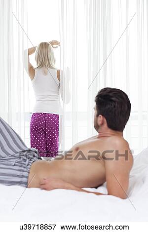 Stock Photo - Shirtless man looking at woman standing by window in hotel room. Fotosearch