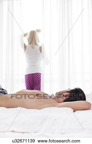 Shirtless young man sleeping in bed with woman standing by window View Large Photo Image Stock Photo - Shirtless young man sleeping in bed with woman standing by window. Fotosearch